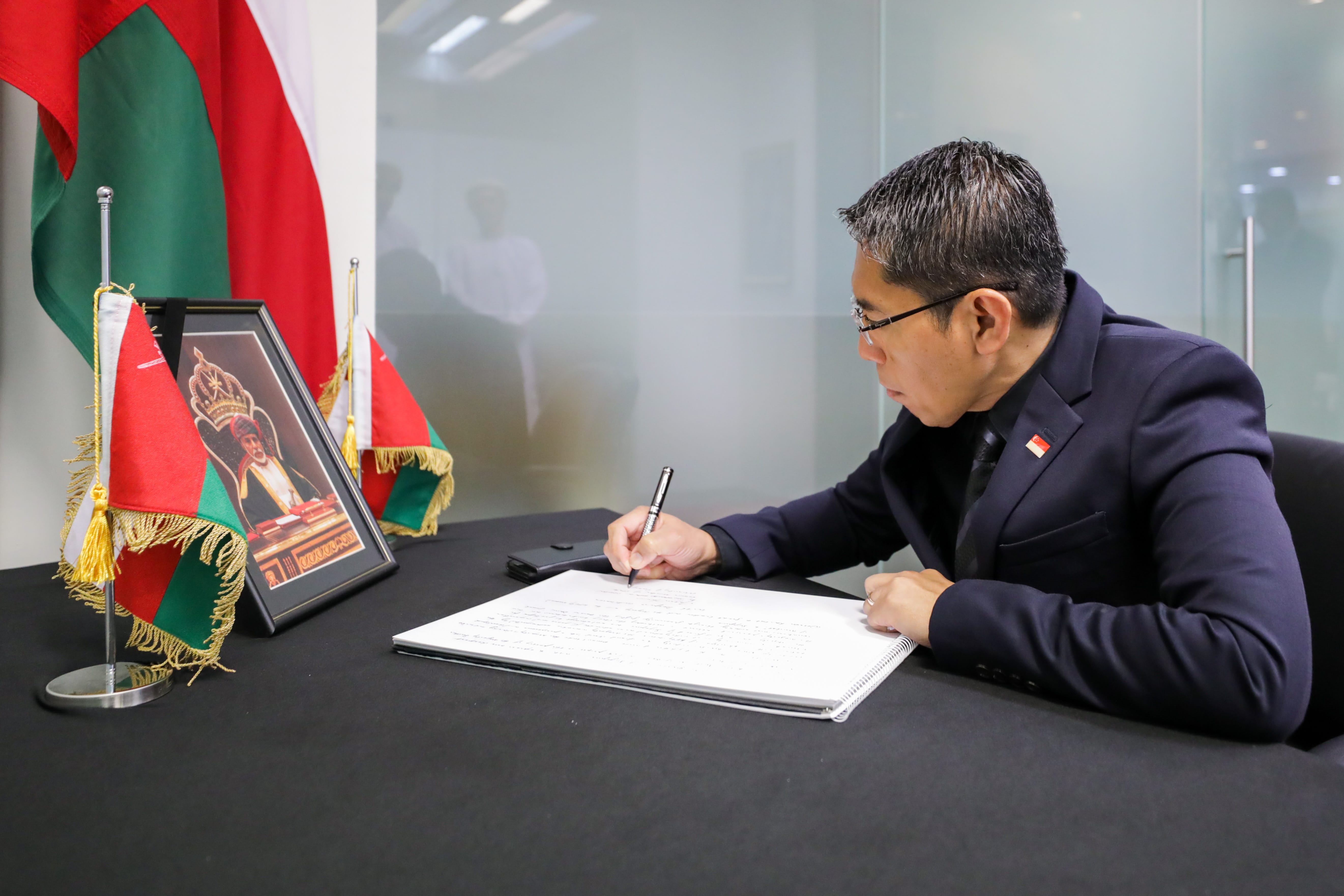 Man in suit signing document at desk with Oman flags and framed portrait nearby.